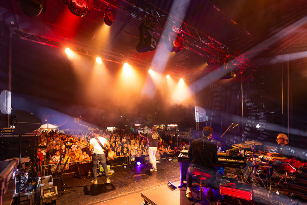 Vue large depuis les backstage d’un concert en plein air, scène éclairée et public rassemblé