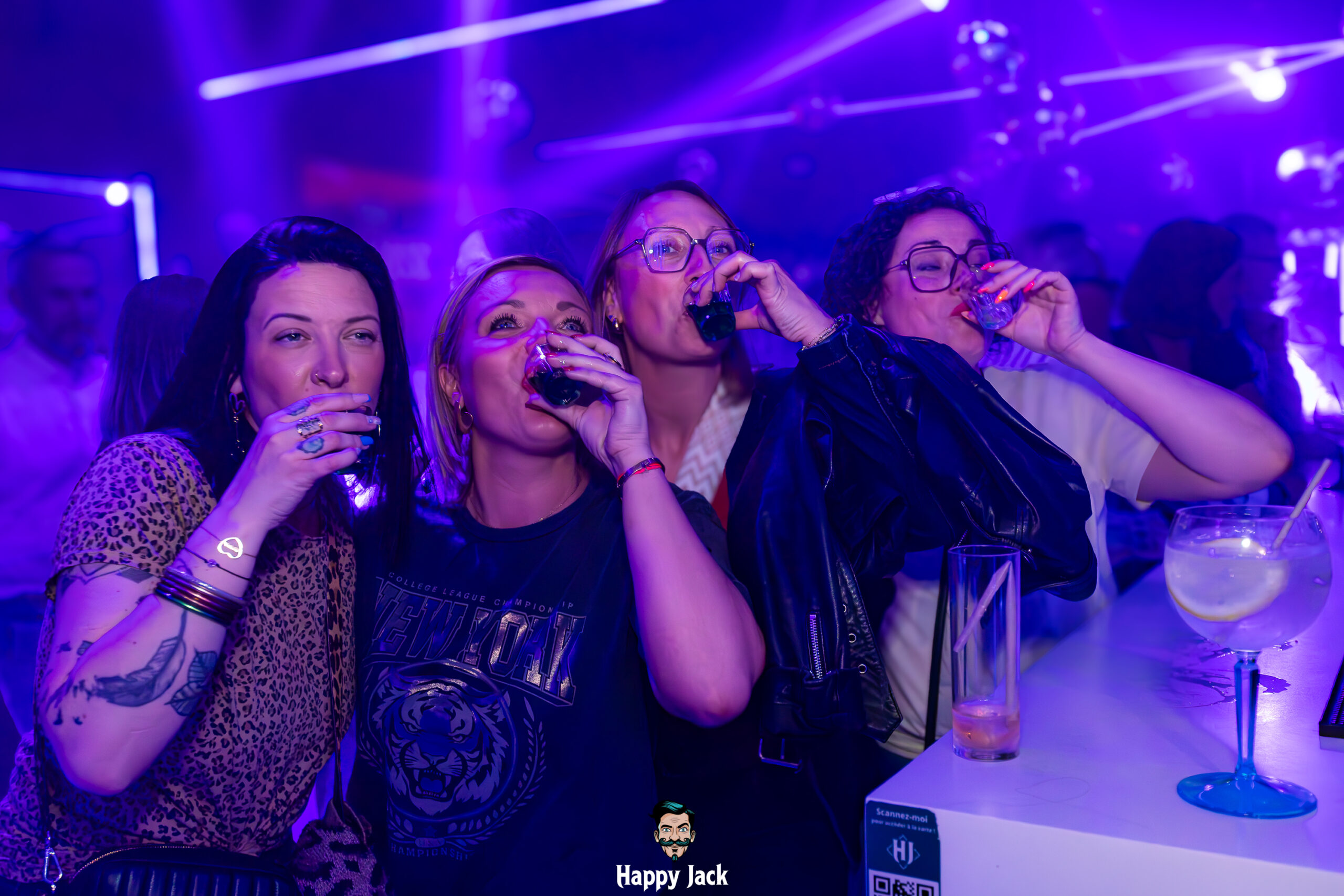 Clients du club prenant des shots en même temps au comptoir du bar sous des lumières néons violettes.