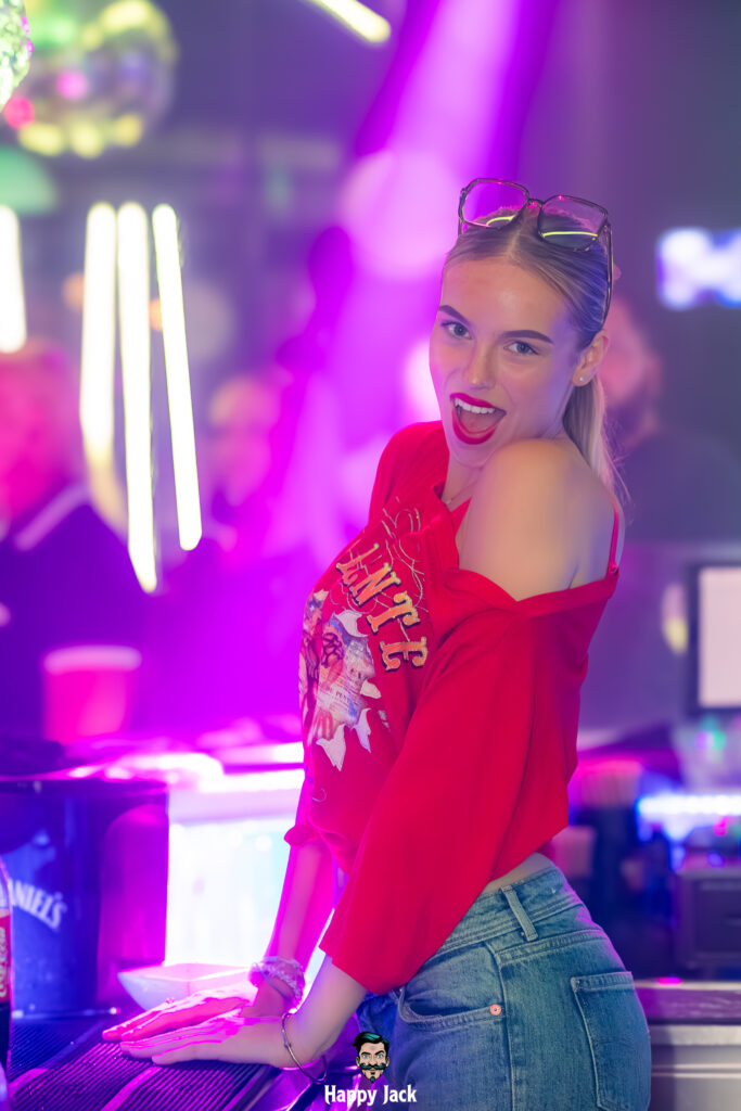 Portrait d'une clubbeuse en t-shirt rouge et jean souriant au bar du club Happy Jack.