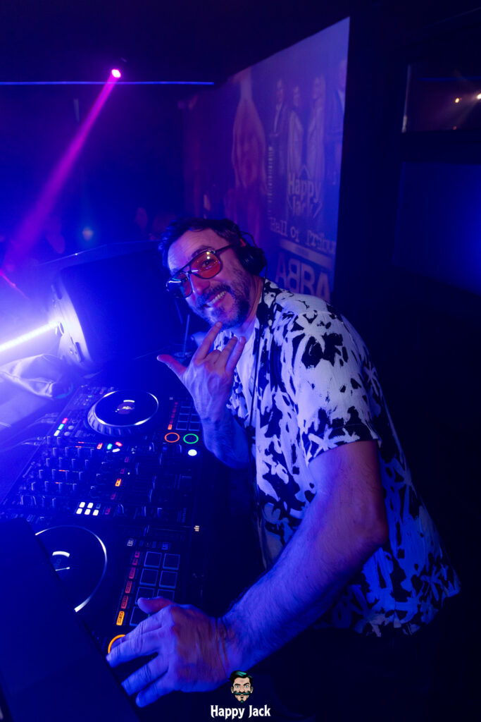 DJ souriant en chemise imprimée faisant un signe de la main derrière sa régie de mixage en boîte de nuit.